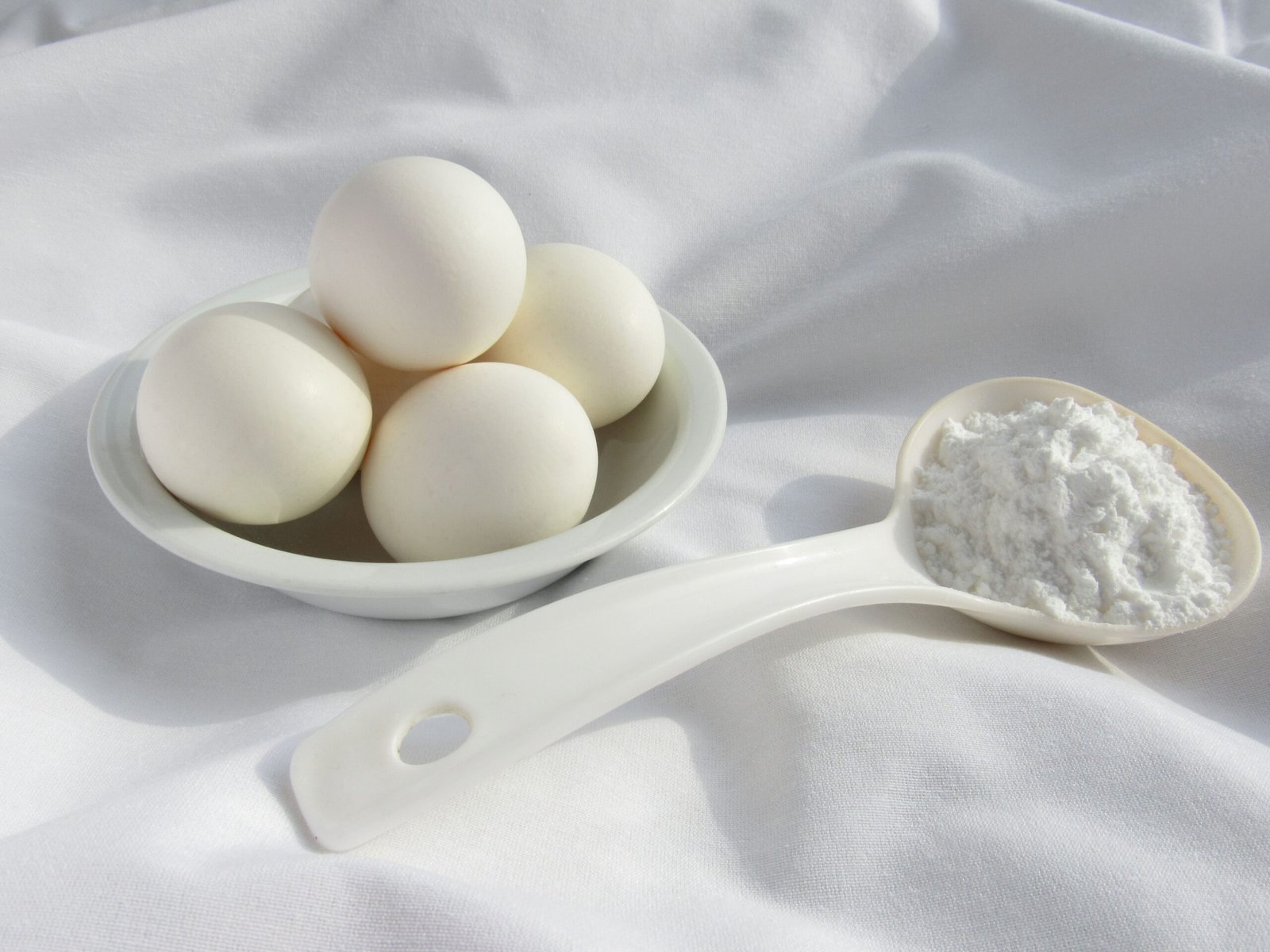 High-angle view of eggs in a bowl and flour in a spoon on a white fabric background.
